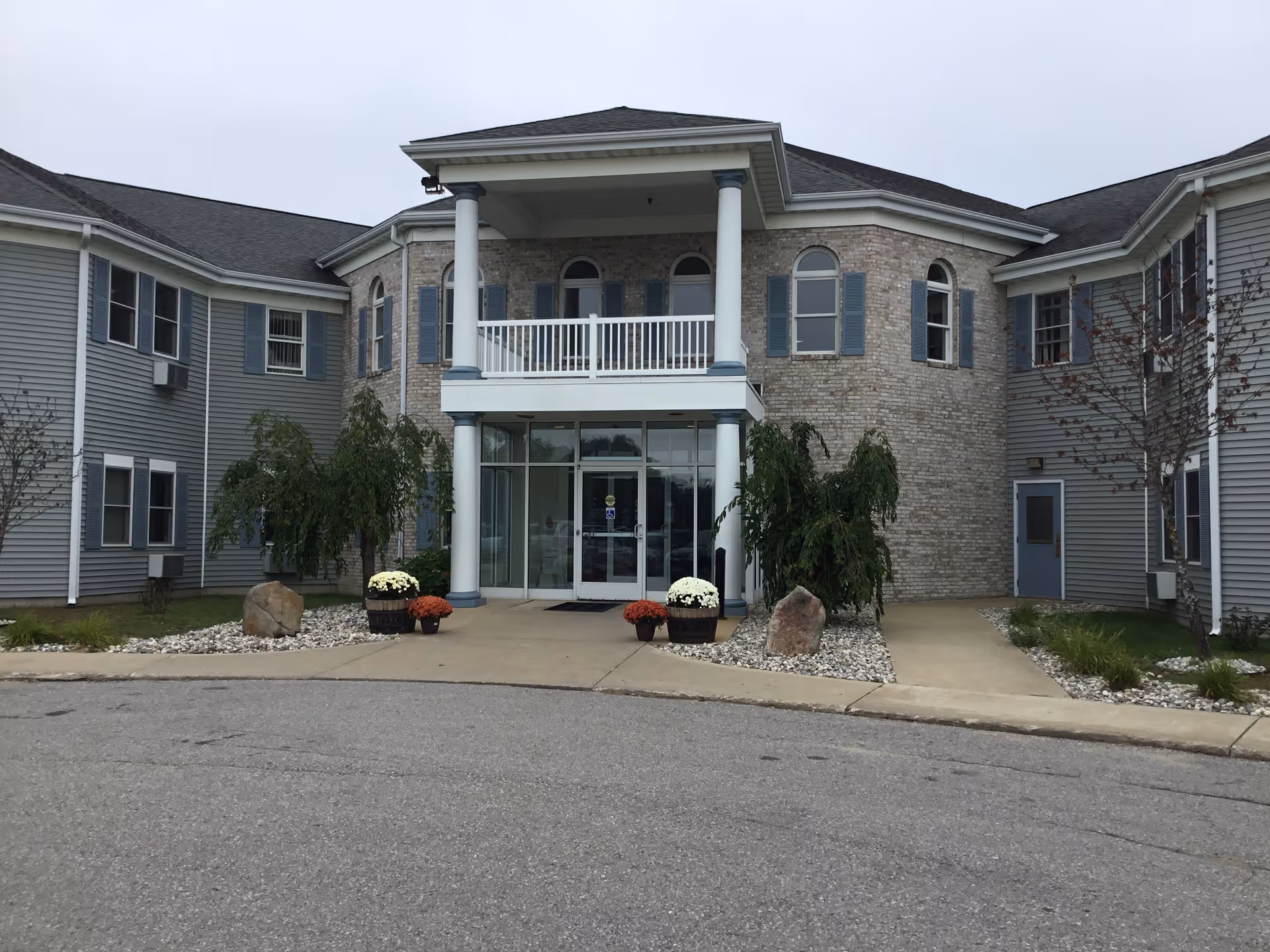 Front exterior view of Whispering Pines Senior Living facility showing a two-story building with a central entrance featuring glass doors, white columns, a balcony above, and landscaping with small trees, rocks, and potted flowers.