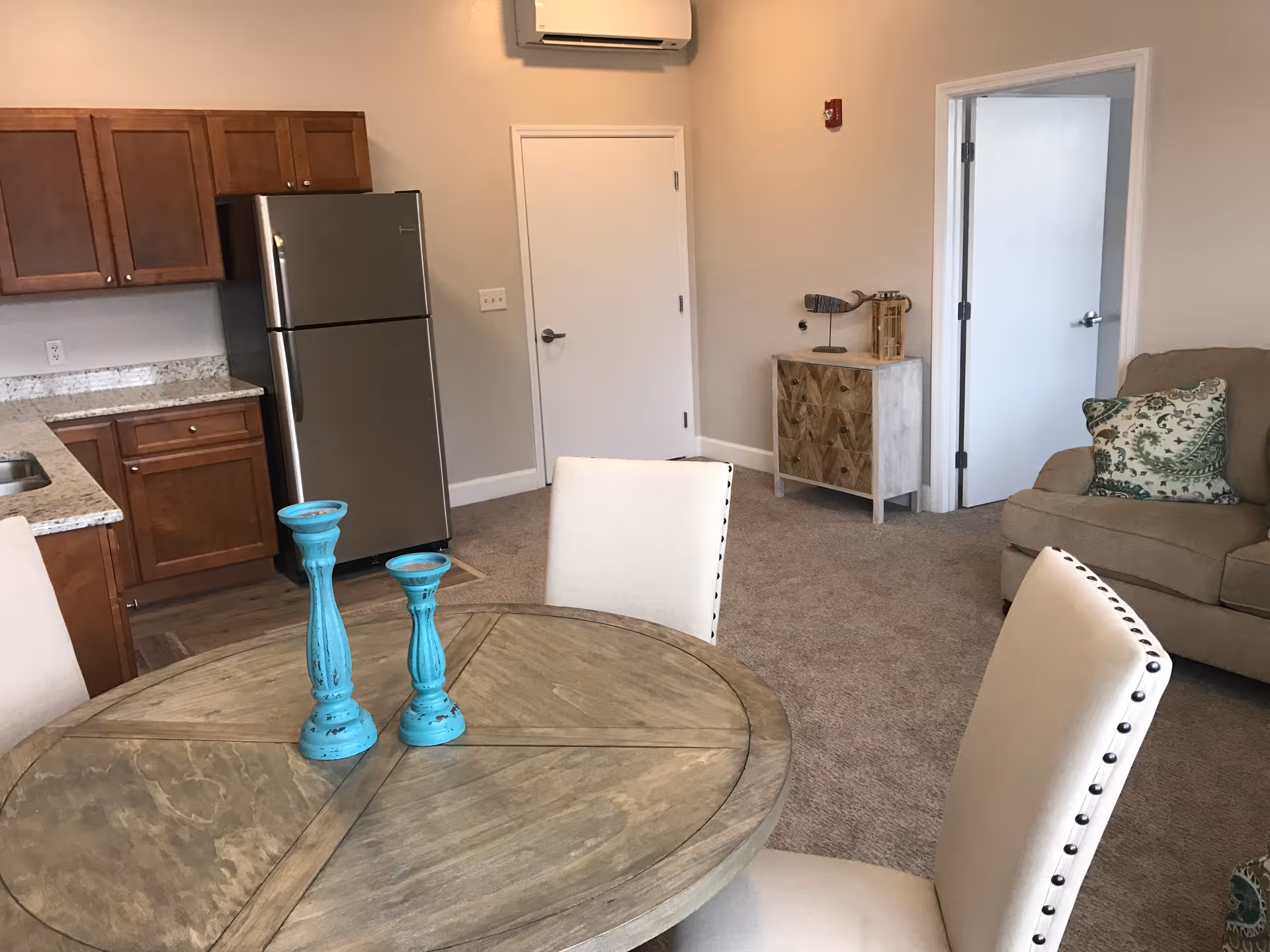 Interior view of a senior living facility showing a small kitchen area with wooden cabinets, a stainless steel refrigerator, and granite countertops. In the foreground, there is a round wooden dining table with two blue decorative candle holders and cream-colored chairs with nailhead trim. To the right, there is a beige sofa with a green patterned pillow and a small wooden cabinet with decorative items on top. The room has beige carpet and light-colored walls with two white doors visible.