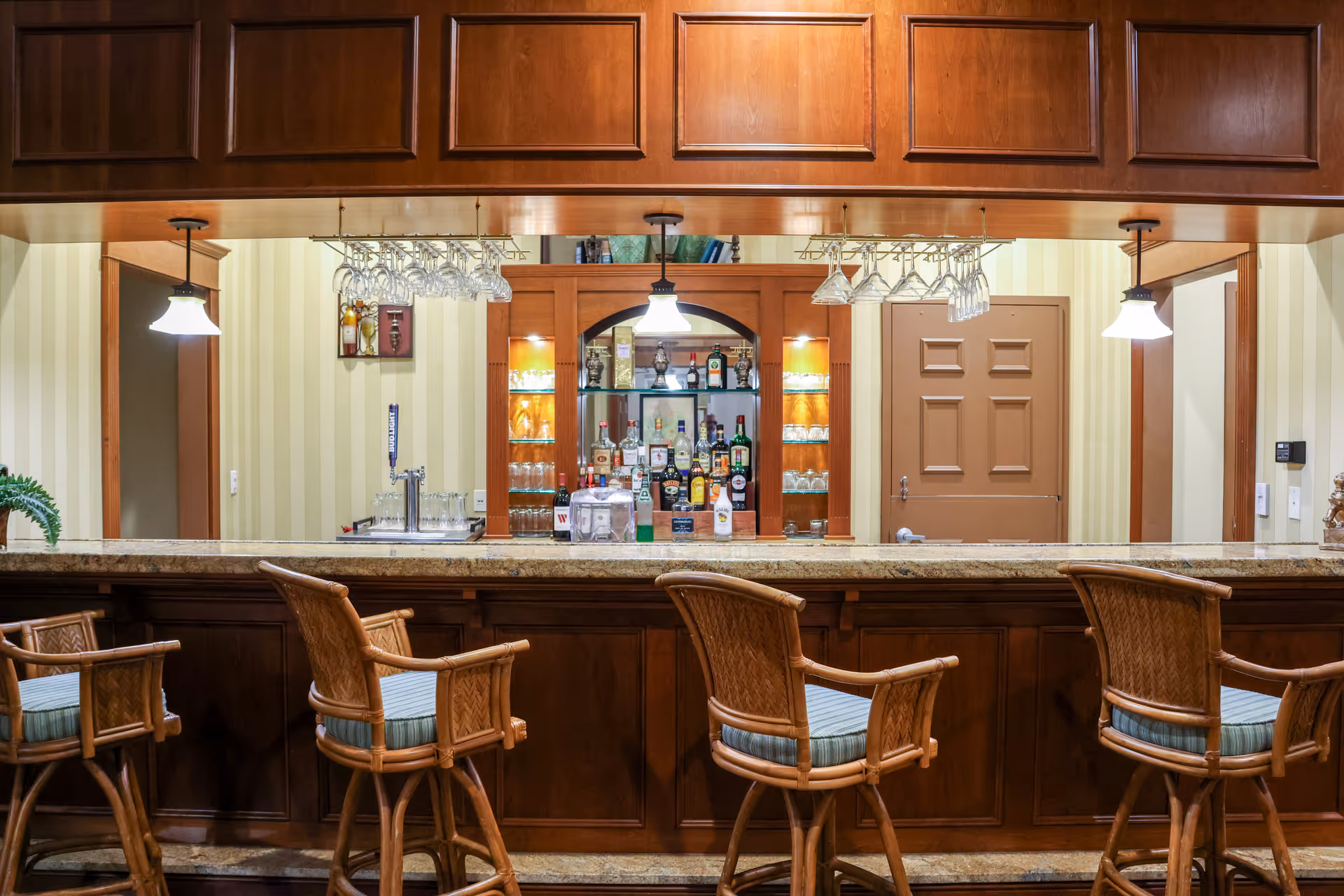 Interior view of a bar area in a senior living facility featuring a wooden counter with four cushioned bar stools. Behind the counter, there is a mirrored shelving unit stocked with various bottles of liquor and glassware. Overhead, hanging racks hold upside-down wine glasses, and three pendant lights illuminate the space. The walls have a striped wallpaper and there are two doorways visible on either side of the shelving unit.