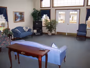 Communal living area with blue sofas, a side table, TV, potted plants, and double doors with transom windows.