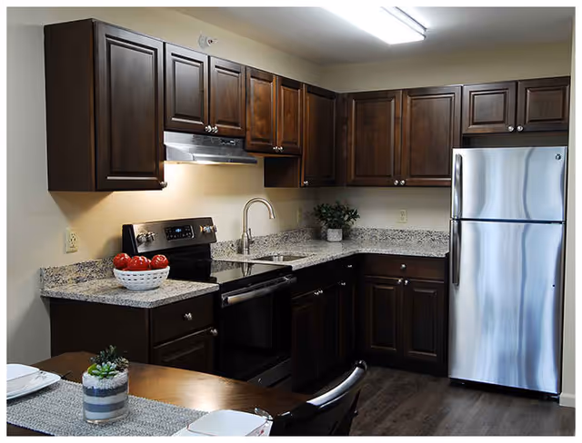 Modern kitchen with dark wooden cabinets, granite countertops, stainless steel refrigerator and stove, a sink with a curved faucet, and a bowl of red apples on the counter. A dining table with place settings and a small plant is partially visible in the foreground.