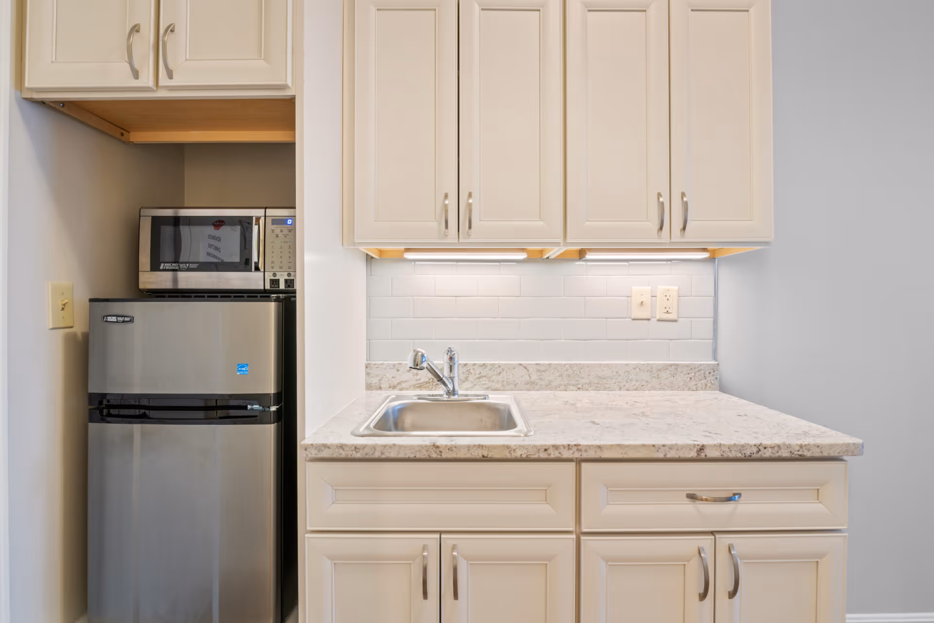 A compact kitchen area featuring a small stainless steel refrigerator with a microwave on top, white cabinetry above and below a marble countertop, and a stainless steel sink with a faucet. The backsplash is made of white subway tiles, and there are electrical outlets on the wall above the countertop.