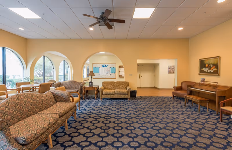 A spacious common area with patterned blue carpet, multiple cushioned sofas and chairs, a wooden piano with a bench, a ceiling fan, and large arched windows letting in natural light. The walls are painted beige and decorated with a framed painting and a bulletin board.