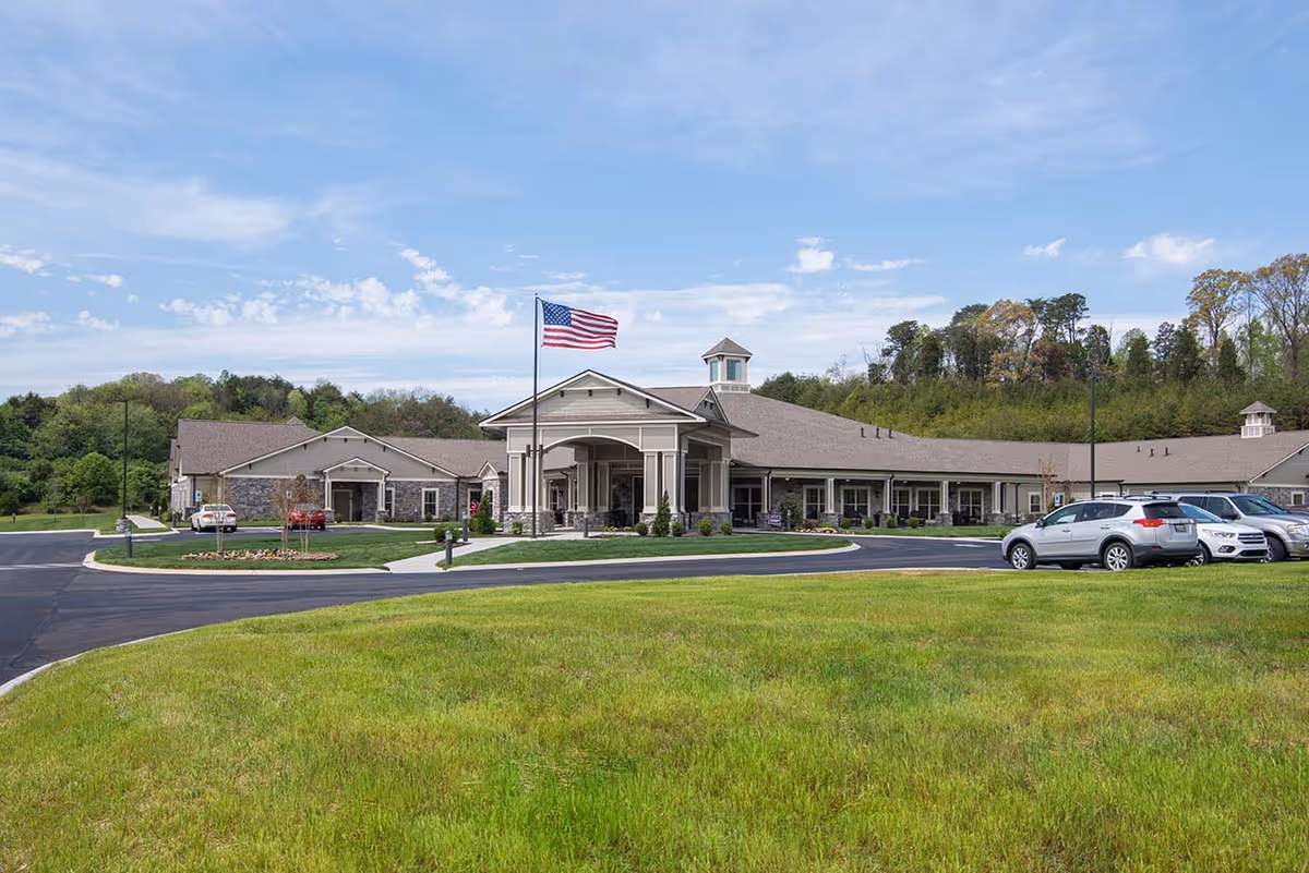 Exterior view of a single-story senior living facility building with a covered entrance, an American flag flying on a flagpole, several parked cars, and a well-maintained lawn in the foreground under a partly cloudy sky.