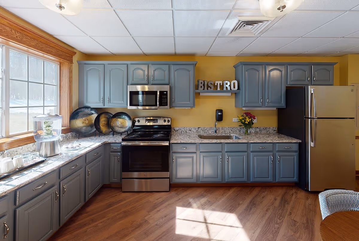 A bright kitchen with blue-gray cabinets, a stainless steel stove and microwave, a stainless steel refrigerator, and a granite countertop. There is a large window on the left letting in natural light. Decorative plates are displayed on the countertop, and a small shelf on the wall holds letters spelling 'BISTRO'. A vase with flowers is placed near the sink.