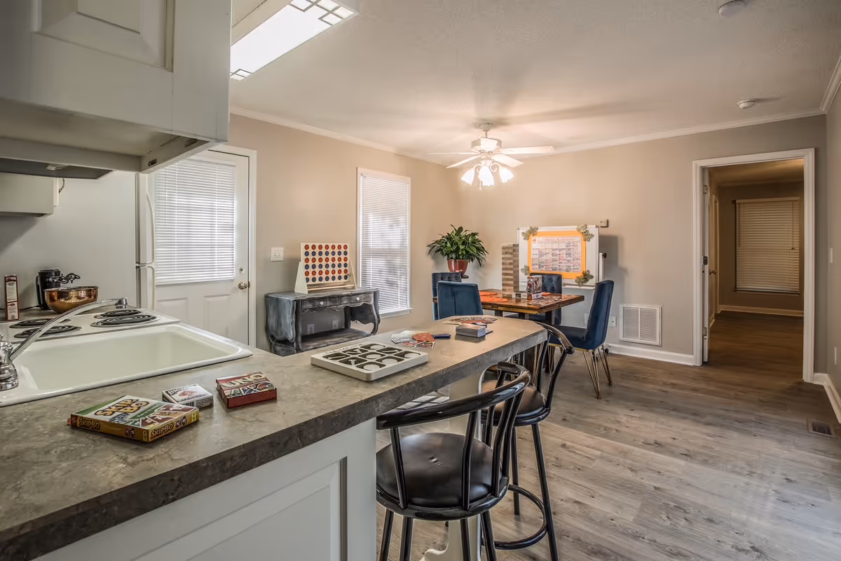 Interior view of a senior living facility's common area featuring a kitchen counter with two black bar stools. On the counter are several card games and a board game. In the background, there is a dining table with four blue chairs, a plant, a large Connect Four game, and a whiteboard with an activity schedule. The room has light-colored walls, wood flooring, and a ceiling fan with lights.