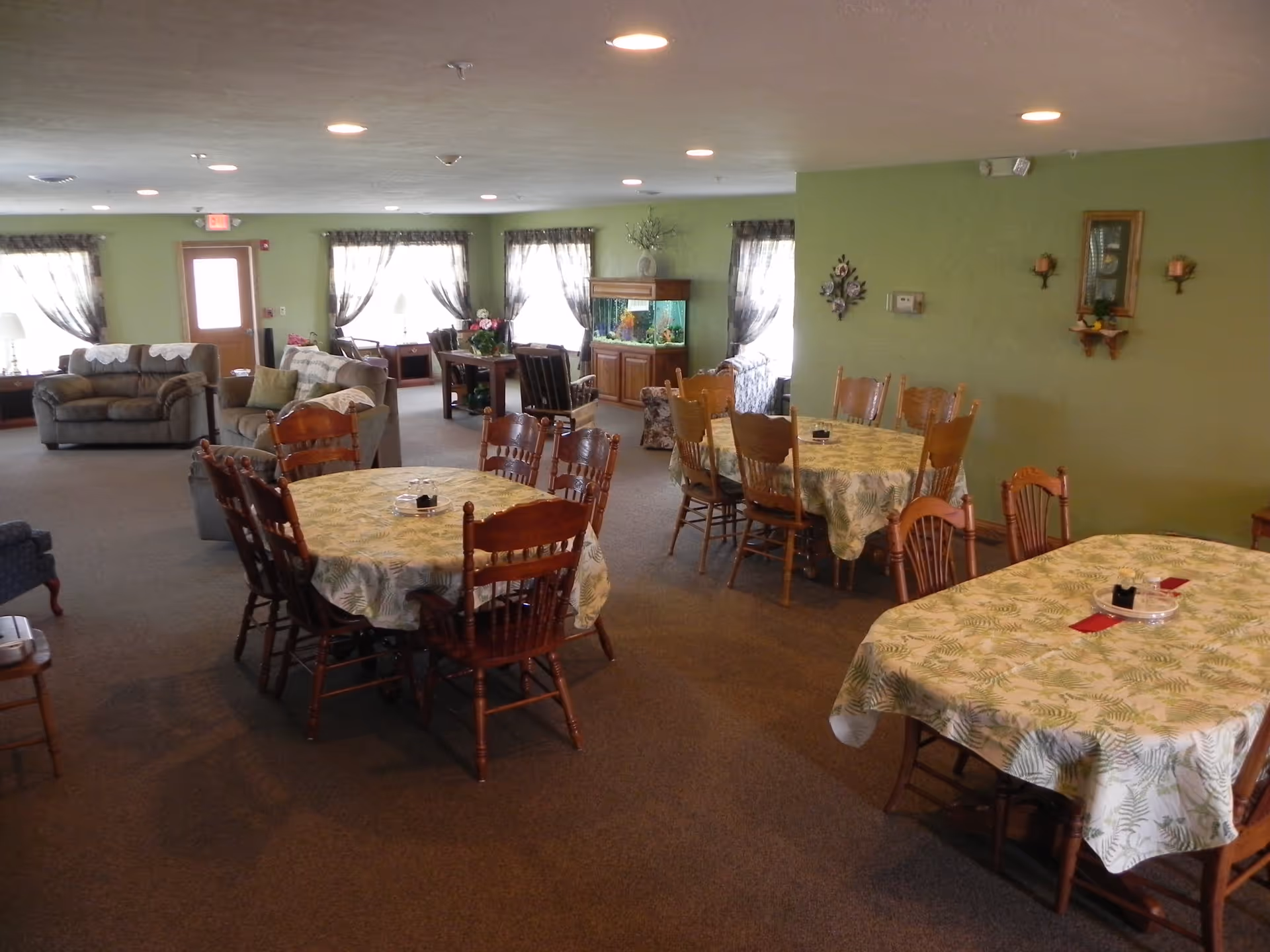 A spacious common area in an assisted living facility with multiple wooden tables covered with green and white patterned tablecloths, surrounded by wooden chairs. In the background, there are several sofas, armchairs, windows with sheer curtains, and a large fish tank against a green wall. The room is well-lit with ceiling lights.