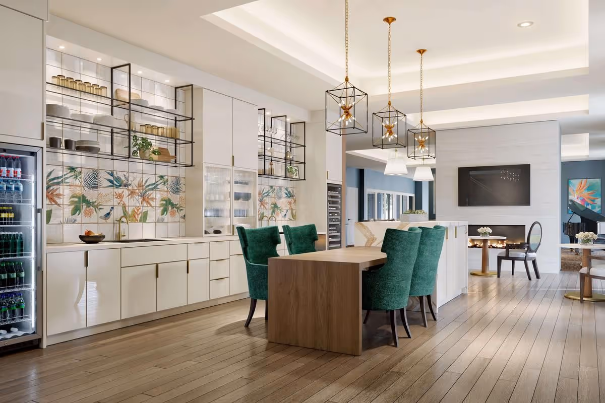 Open-plan communal dining and lounge area with a kitchen counter, green upholstered chairs around a wooden table, decorative pendant lights, and a fireplace with a TV in the background.