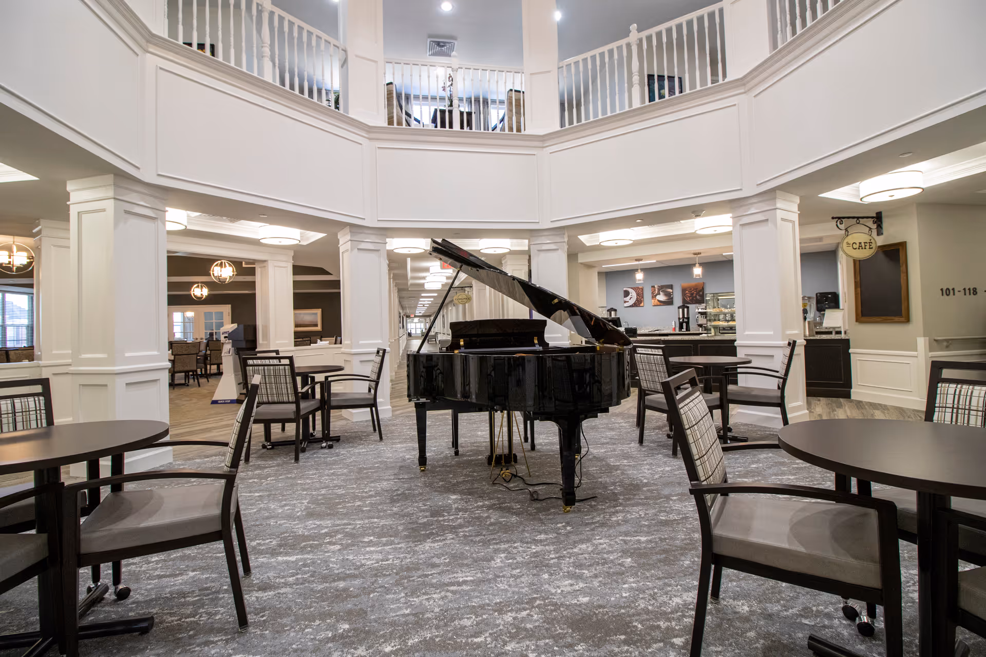 Interior view of a senior living facility common area with a black grand piano in the center, surrounded by round tables and chairs. The space features white pillars and railings on the upper level, with a cafe area visible in the background.