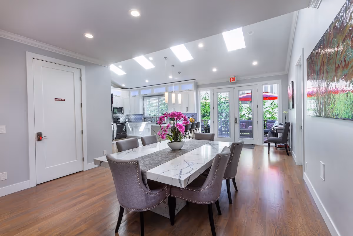 A modern dining area with a rectangular marble table surrounded by six upholstered chairs. A vase with vibrant pink flowers is centered on the table. The room has wooden flooring and white walls, with a kitchen visible in the background featuring white cabinets and stainless steel appliances. Skylights and recessed lighting illuminate the space. Glass doors at the far end open to an outdoor patio with red umbrellas and greenery.
