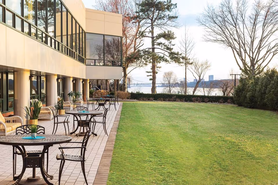 Outdoor patio area at RiverSpring Assisted Living with several metal tables and chairs arranged on a tiled surface next to a well-maintained grassy lawn. The building has large windows and columns, and trees and a body of water are visible in the background under a partly cloudy sky.