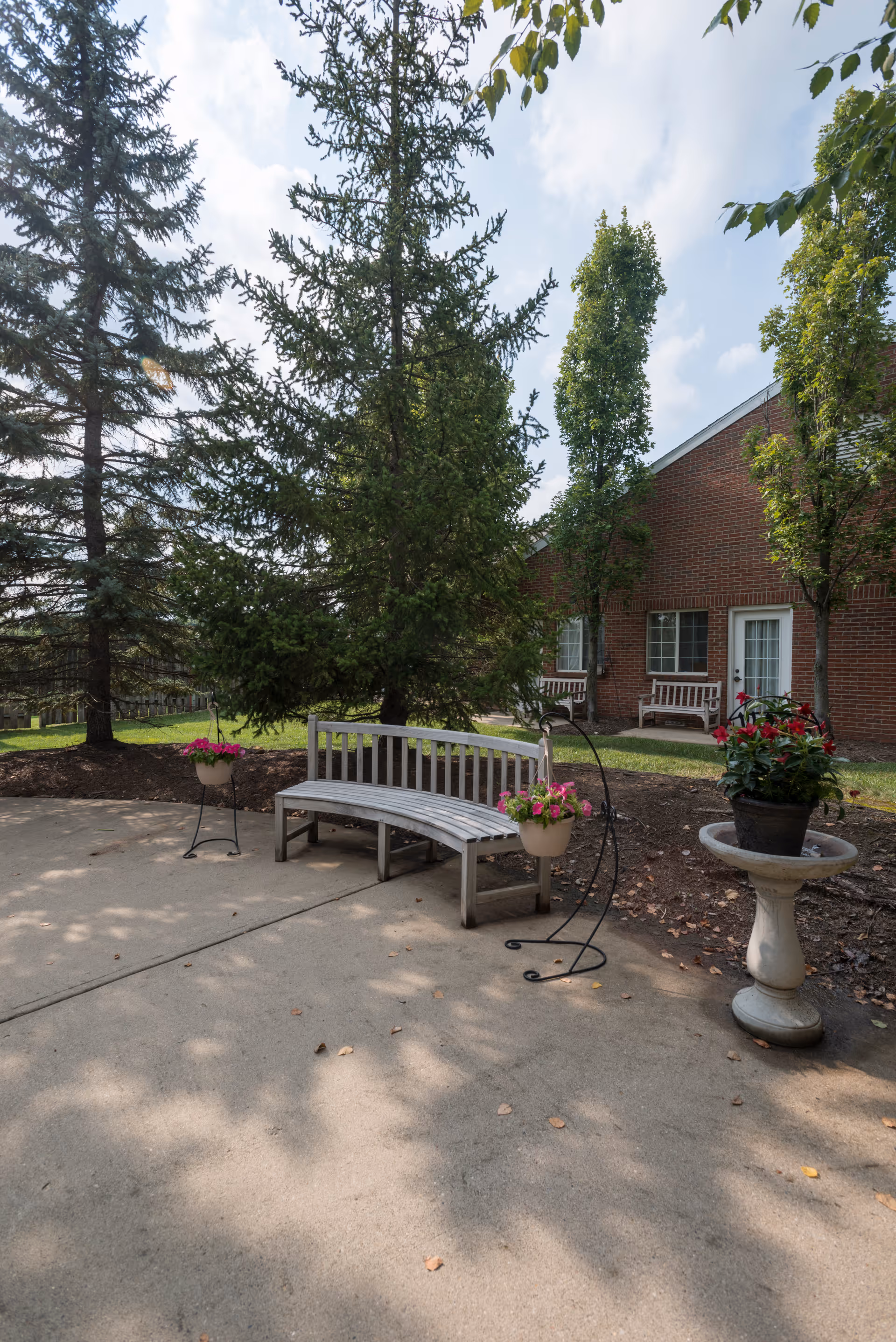 Outdoor patio area with a curved wooden bench, two hanging flower pots with pink flowers, a large potted plant on a pedestal, and several tall trees. In the background, there is a brick building with windows and white benches near the entrance.
