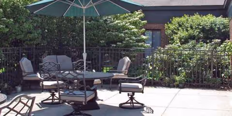 Outdoor patio with table, chairs, umbrella, railing, and surrounding greenery in front of a brick building.
