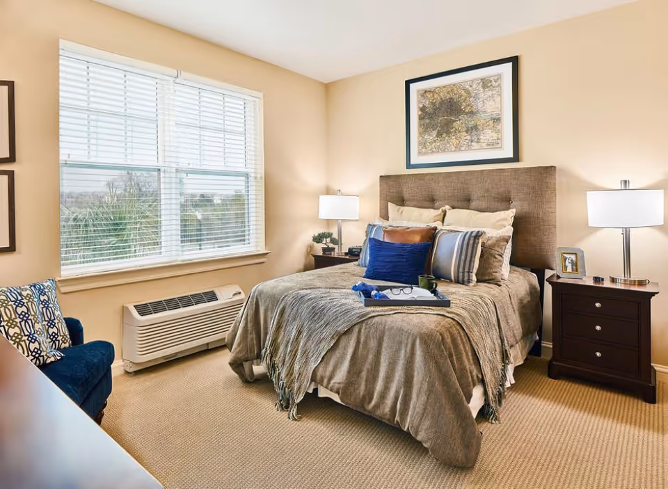 Bright furnished bedroom with a made bed, nightstands and lamps, a window with blinds and an upholstered chair.