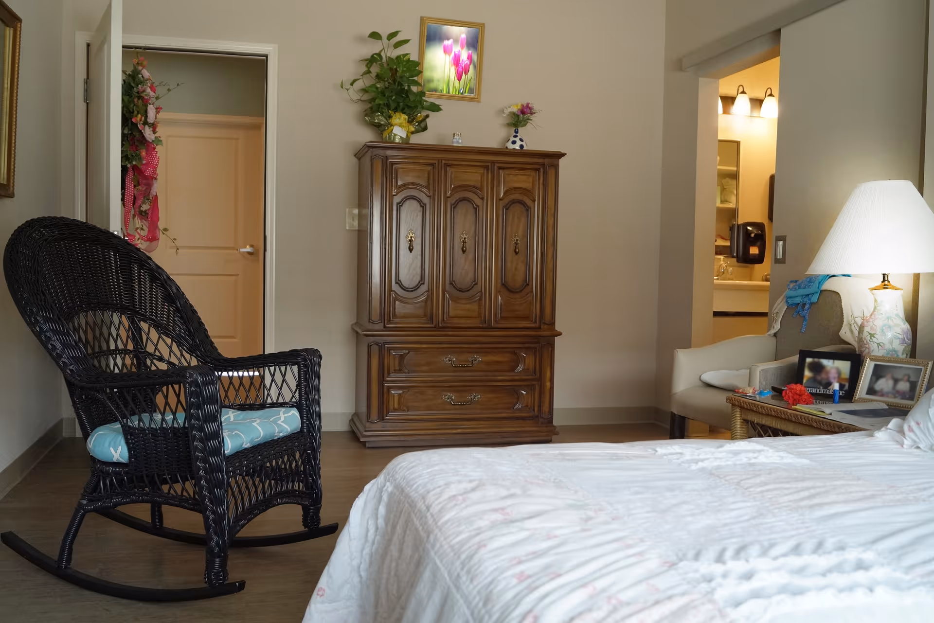A cozy bedroom featuring a black wicker rocking chair with a blue cushion, a wooden armoire with decorative plants and a framed picture on top, a bed with a white comforter, a side table with a lamp, framed photos, and a chair with a blue throw draped over it. The room has beige walls and a door leading to another room.