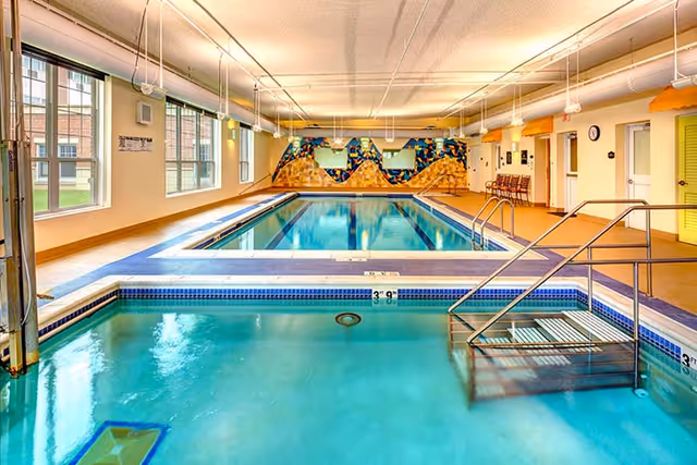 Indoor swimming pool area with a smaller pool in the foreground and a larger pool in the background. The room has large windows on the left side allowing natural light to enter, colorful wall art at the far end, and pool safety equipment including handrails and a pool lift.