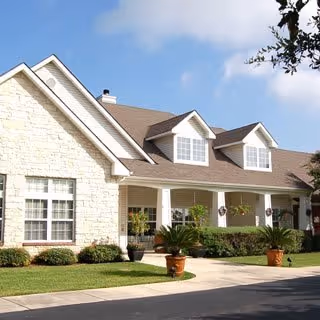 Exterior view of a single-story residential building with stone and siding facade, featuring a covered porch with columns, potted plants, and a well-maintained lawn under a blue sky with some clouds.