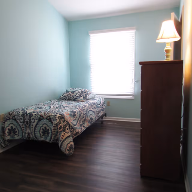 A small bedroom with light blue walls, a single bed with a patterned bedspread and pillow, a window with white blinds, and a wooden dresser with a lit lamp on top.
