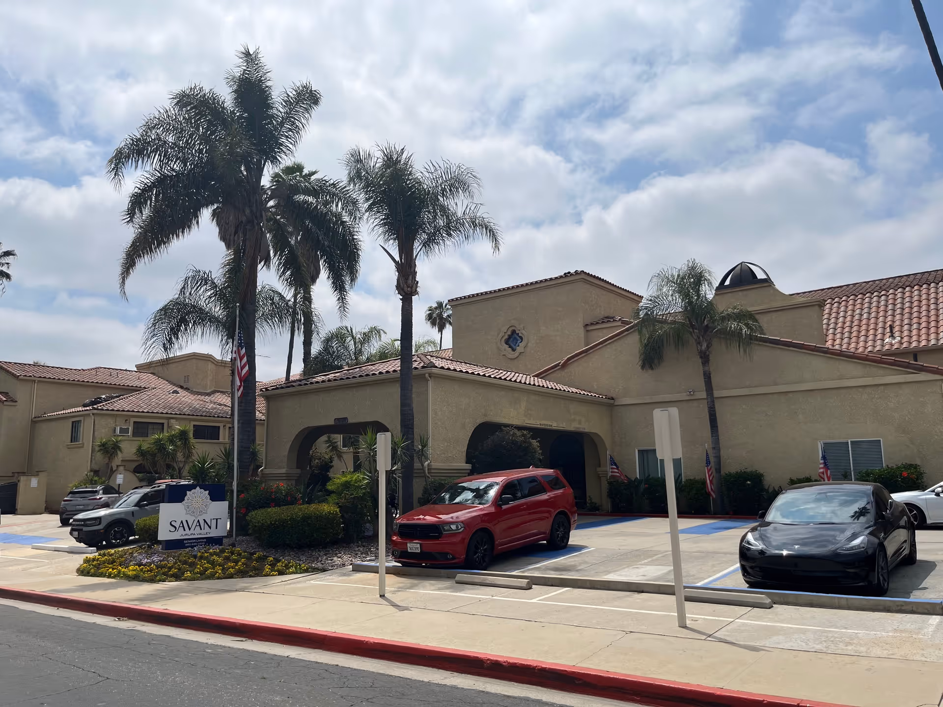 Exterior view of Savant of Jurupa Valley senior living facility showing a beige stucco building with red tile roof, palm trees, parked cars, and a sign with the facility name in front.