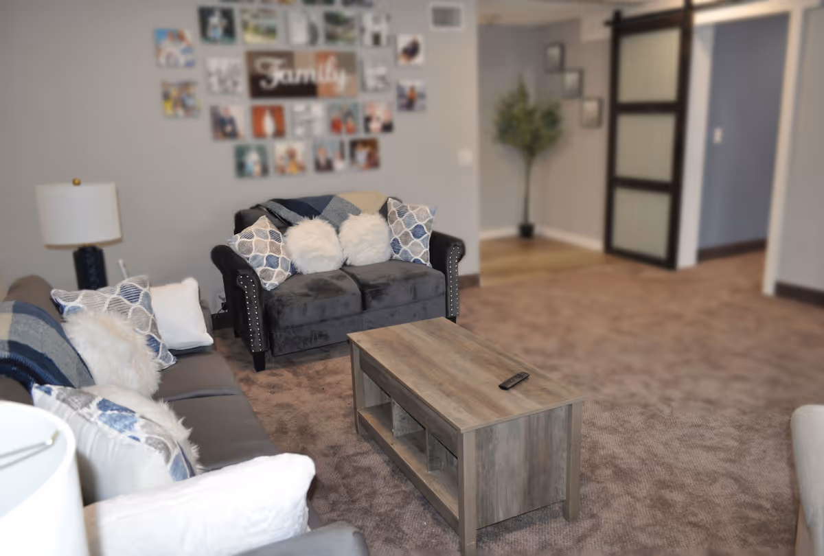 A cozy living room with two sofas adorned with patterned and fluffy pillows, a wooden coffee table with a remote control on it, a lamp on a side table, a wall decorated with family photos and the word 'Family', and a potted plant in the corner near a sliding door.