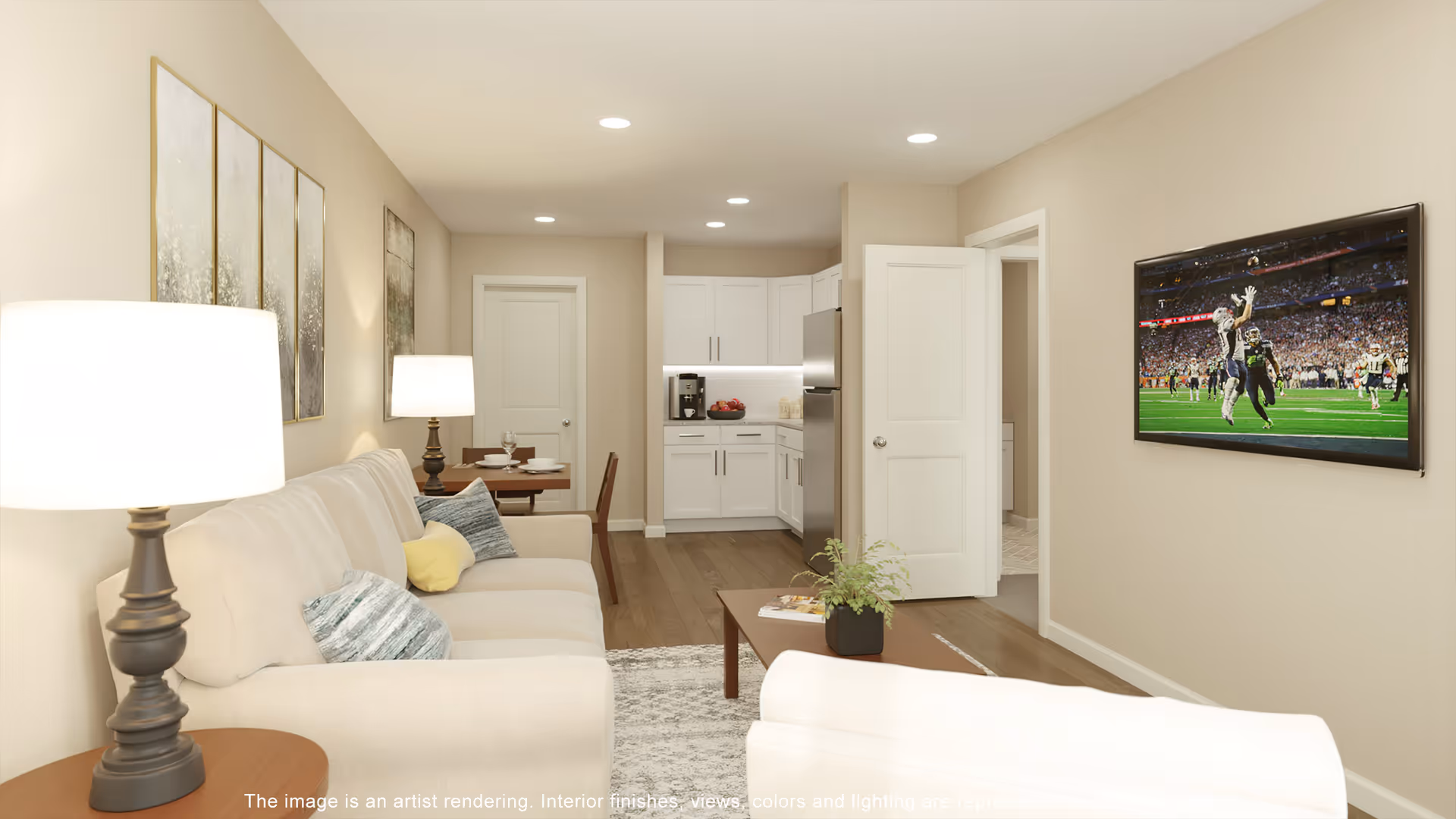 A cozy living room with a beige sofa adorned with blue and yellow pillows, a wooden coffee table with a small plant, and a wall-mounted TV displaying a football game. The room has warm lighting from two table lamps and features light-colored walls and wooden flooring. In the background, there is a small dining area with a table and chairs, and a compact kitchen with white cabinets and stainless steel appliances.