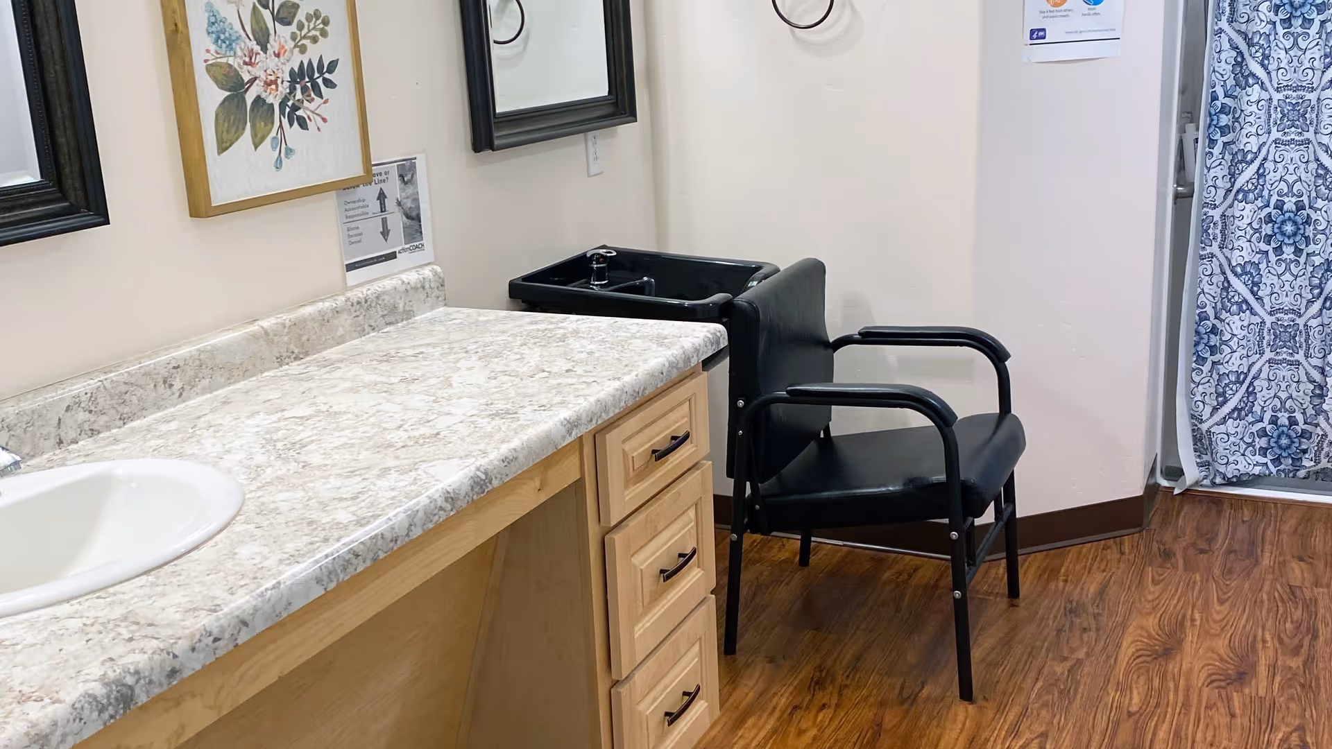Interior view of a room with a countertop and sink on the left, a black salon chair with a black wash basin behind it, wooden flooring, and a blue and white patterned curtain on the right. There are framed pictures and a mirror on the wall above the countertop.