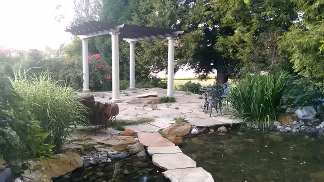 A serene outdoor garden area with a stone pathway crossing a small pond, leading to a pergola with white columns. There are lush green plants, bushes, and trees surrounding the area, along with a metal bench near the pond.