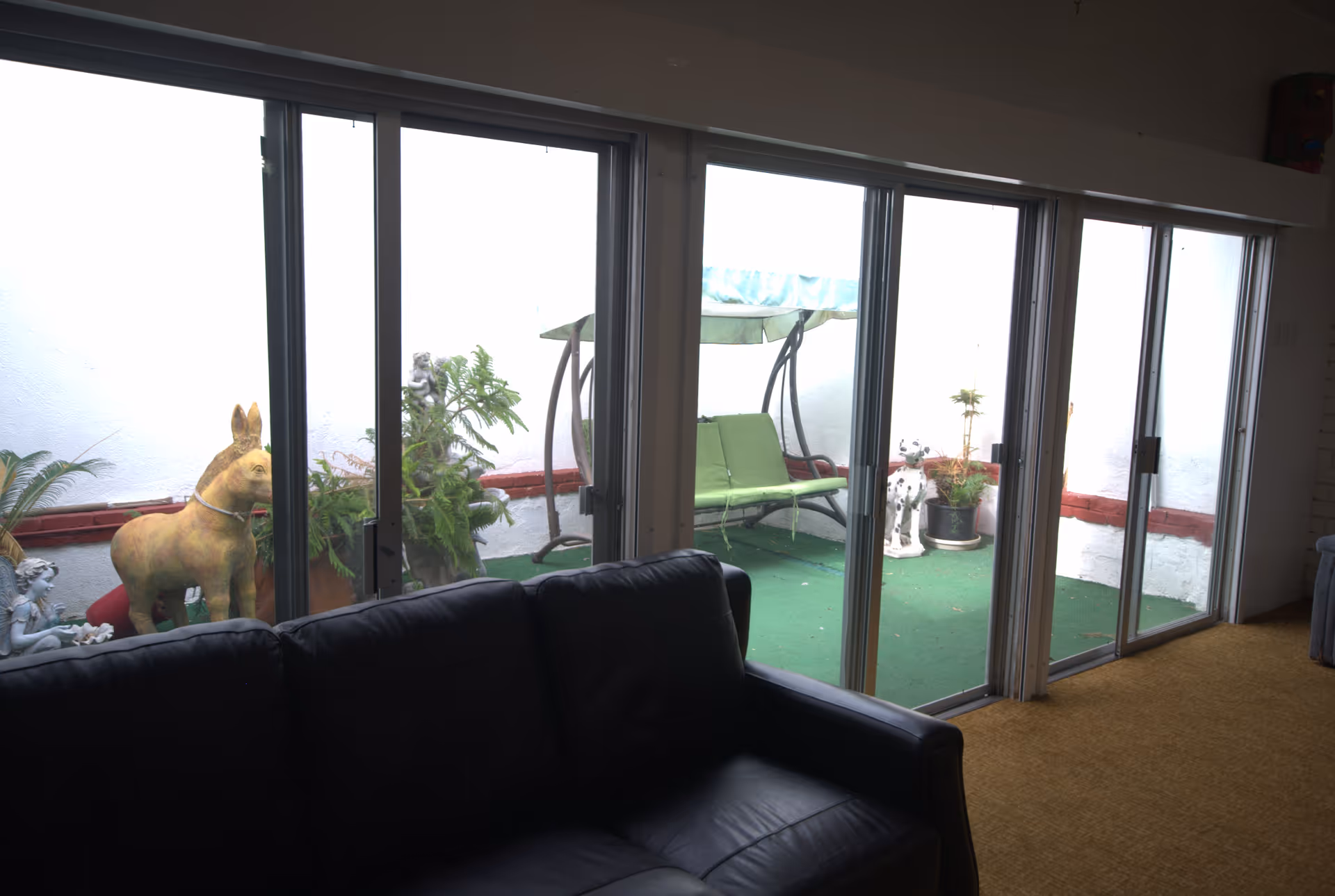 A black leather couch in front of large sliding glass doors that open to a small outdoor patio area with green carpet, potted plants, a green cushioned swing, and decorative statues including a donkey and a small angel.