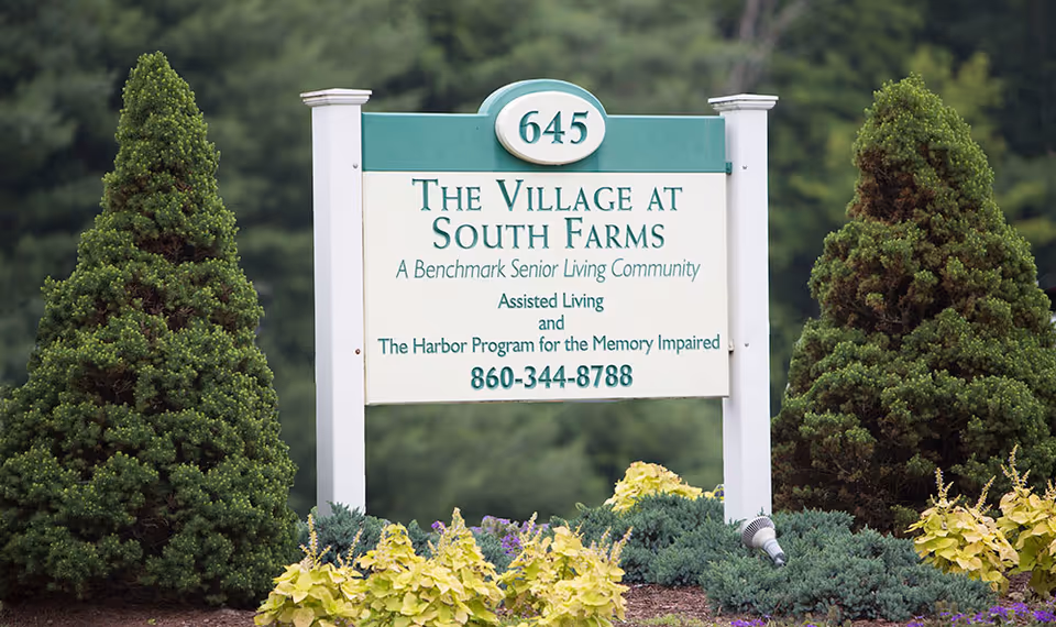 Outdoor sign for The Village at South Farms, a Benchmark Senior Living Community offering assisted living and a memory impaired program, surrounded by greenery and landscaping.