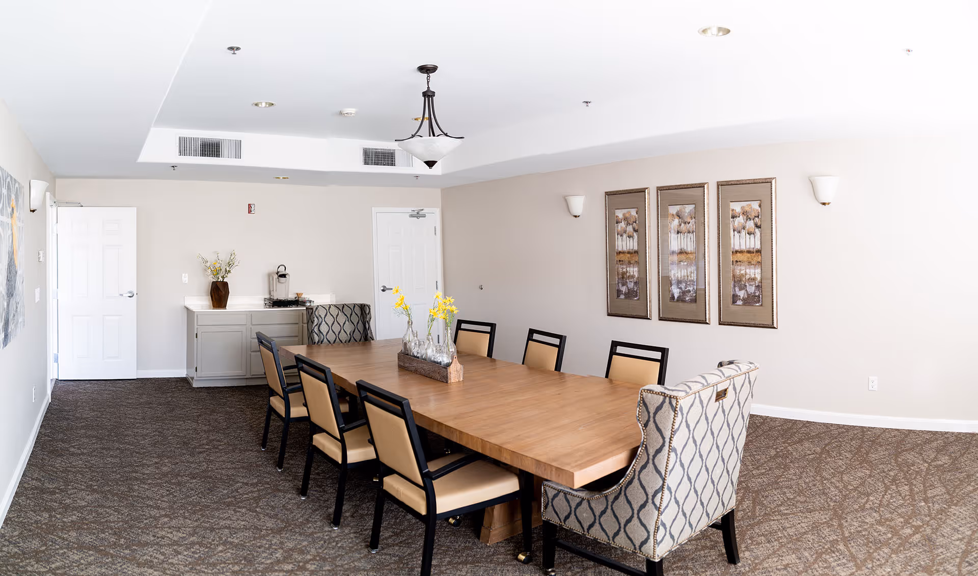 Communal dining room with a long wooden table, upholstered chairs, wall art, and a sideboard.