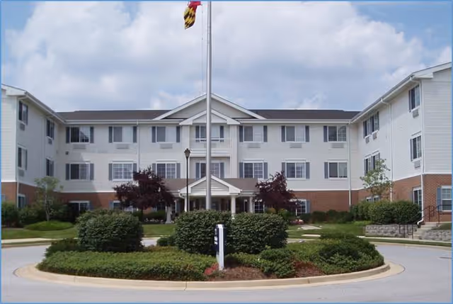 Front exterior view of a three-story senior living facility building with white siding and red brick accents. There is a circular driveway with landscaped bushes and a flagpole with a flag in the center. The sky is partly cloudy.