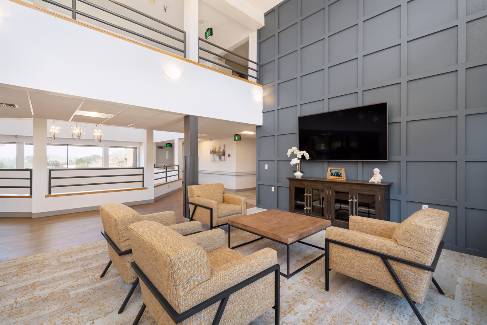 Bright two-story common lounge with four armchairs around a coffee table facing a wall-mounted TV and console.