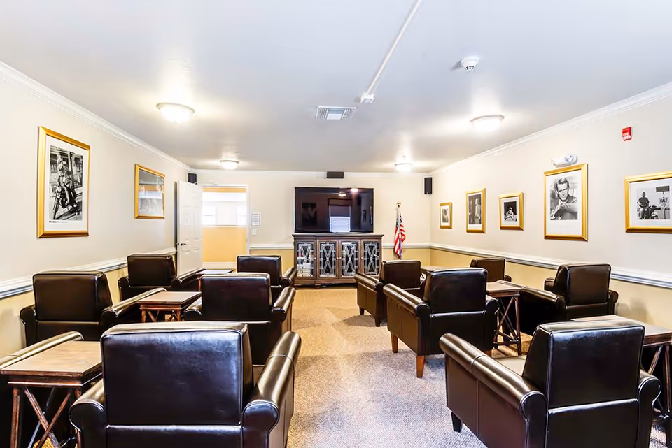 A well-lit common room with multiple black leather armchairs arranged in rows facing a large flat-screen TV mounted on a wooden cabinet. The walls are decorated with framed black and white photographs, and an American flag stands in the corner.