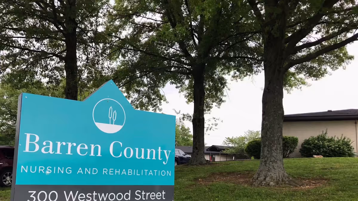 A turquoise sign reading "Barren County Nursing and Rehabilitation" on a grassy lawn with trees and a low building in the background.