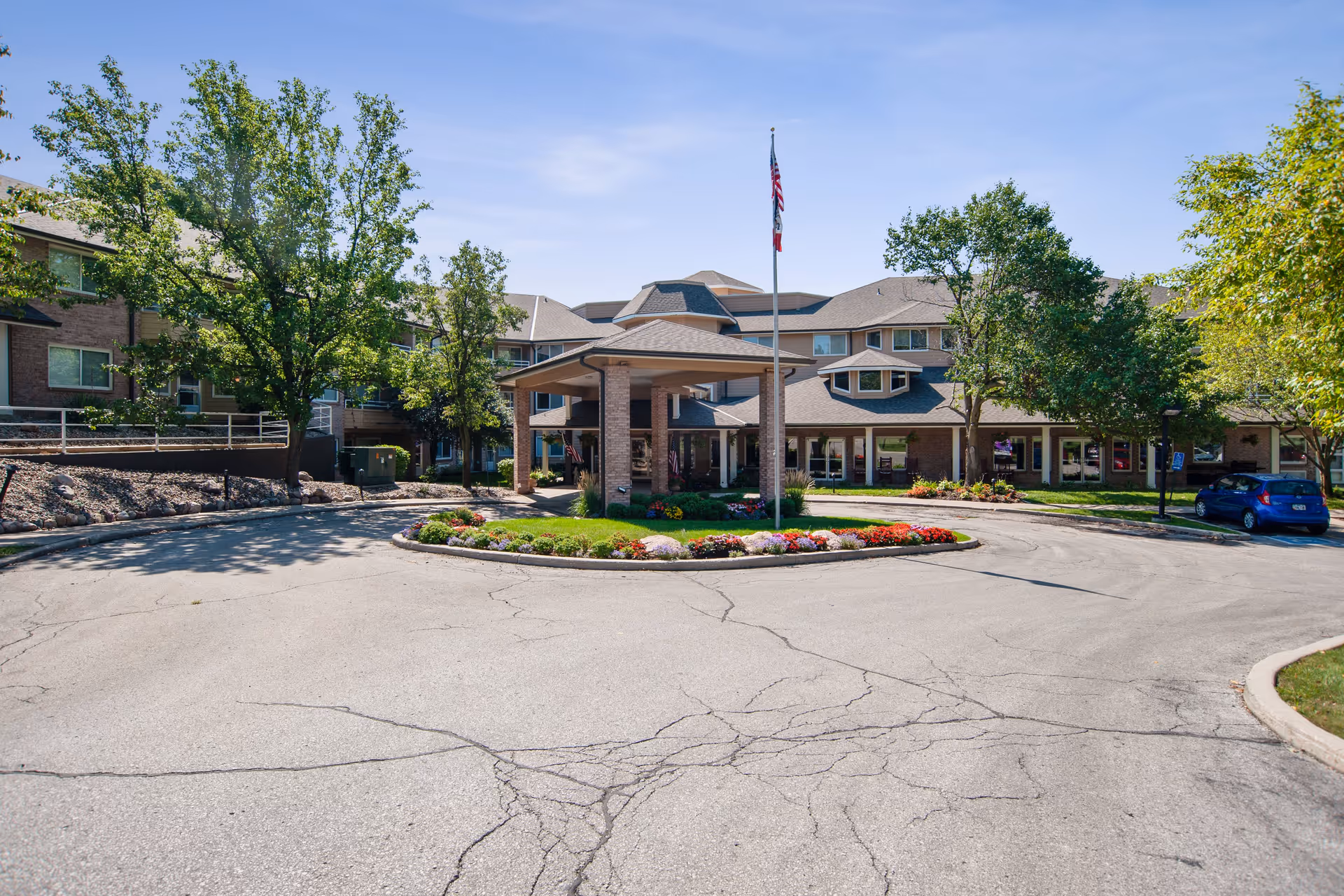 Front exterior view of Independence Village of Des Moines, showing a circular driveway with a landscaped island featuring flowers and a flagpole with an American flag. The building is a multi-story structure with large windows and surrounded by trees under a clear blue sky.