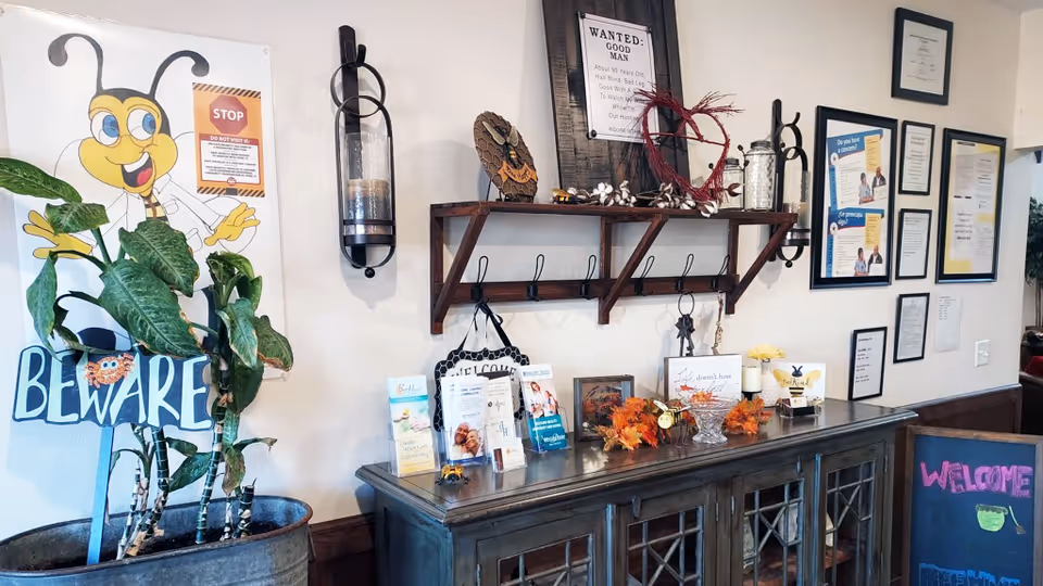 Interior view of a welcoming area in a senior living facility with a large potted plant, a cheerful bee-themed 'Beware' sign, a wooden shelf with hooks holding keys and decorative items, a cabinet with brochures and autumn decorations, framed certificates and informational posters on the wall, and a chalkboard sign that says 'Welcome'.