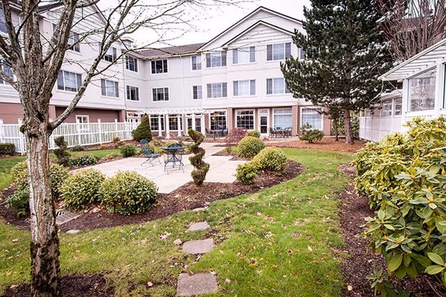 Courtyard garden with a small paved patio, chairs and trimmed shrubs in front of a multi-story senior living building.