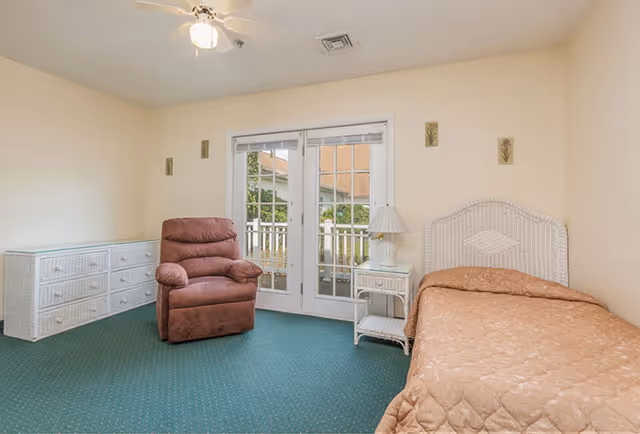 A bedroom with a single bed covered in a brown quilt, a white wicker nightstand with a lamp, a brown recliner chair, a white wicker dresser, and double glass doors leading to an outdoor area. The room has beige walls and green carpet.