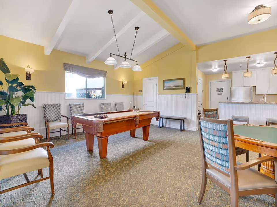 A well-lit recreational room with a pool table in the center, surrounded by chairs along the walls. There is a large window with a curtain, a potted plant, and a card table with chairs in the foreground. The room has yellow walls with white wainscoting and a carpeted floor. In the background, there is a kitchen area with white cabinets and pendant lights.