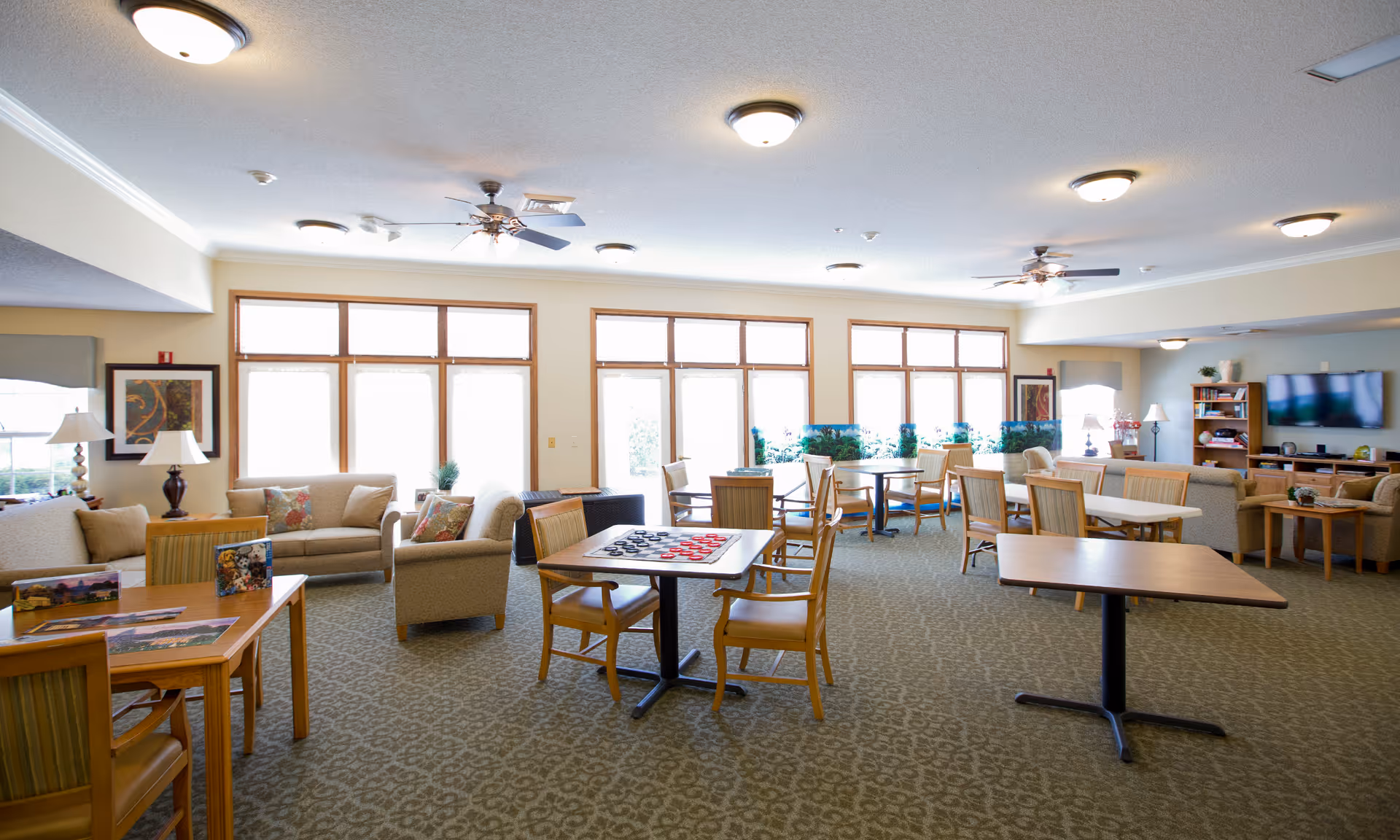 A spacious, well-lit common area in a senior living facility with multiple seating arrangements including sofas, armchairs, and tables with chairs. The room has large windows letting in natural light, ceiling fans, and a television mounted on the wall. One table has a checkers game set up.