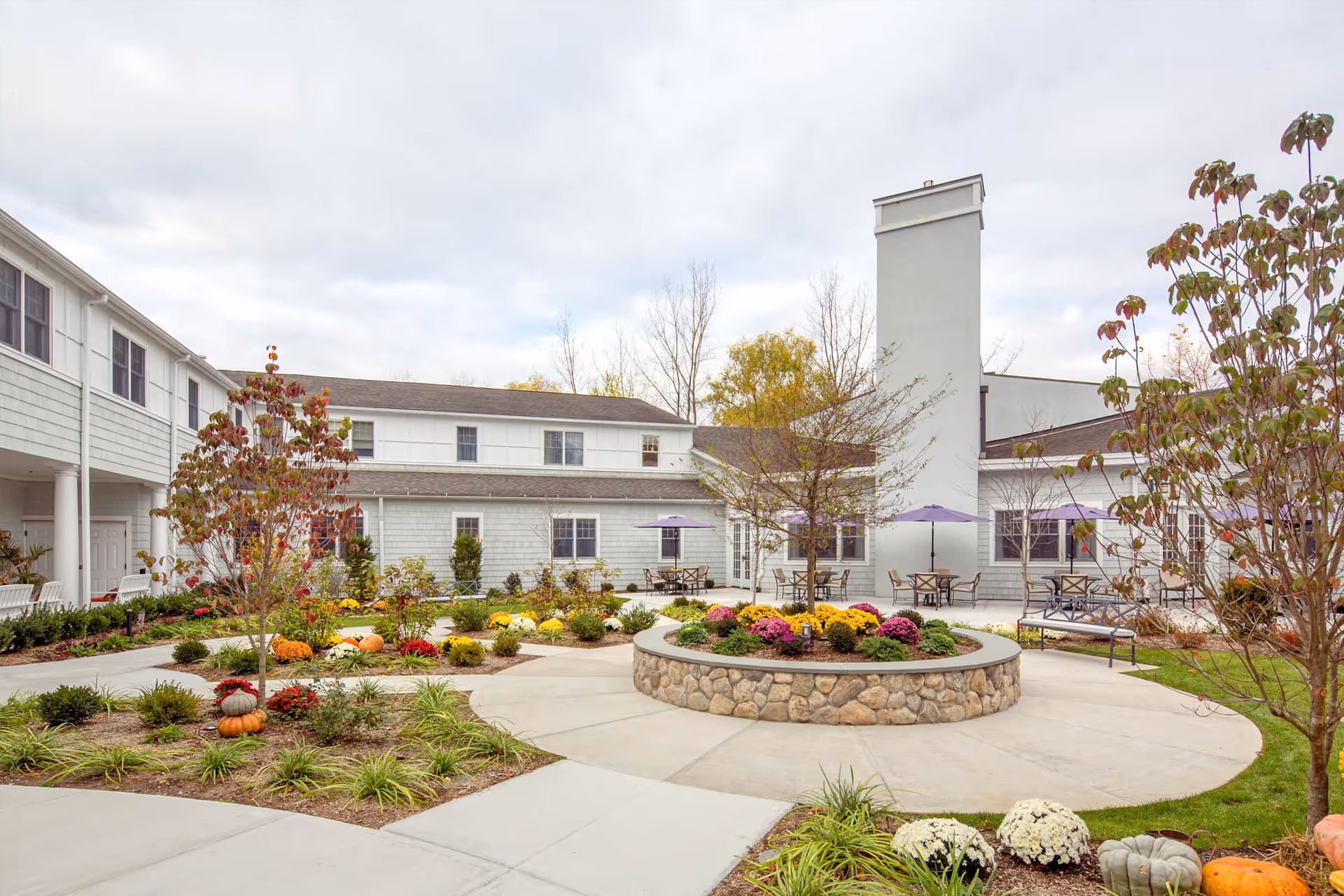 Outdoor courtyard area of a senior living facility with a circular stone planter filled with colorful flowers in the center. Surrounding the planter are paved walkways, small trees, and landscaped garden beds with pumpkins and plants. There are tables with purple umbrellas and chairs near the building, which is a two-story structure with white siding and multiple windows.