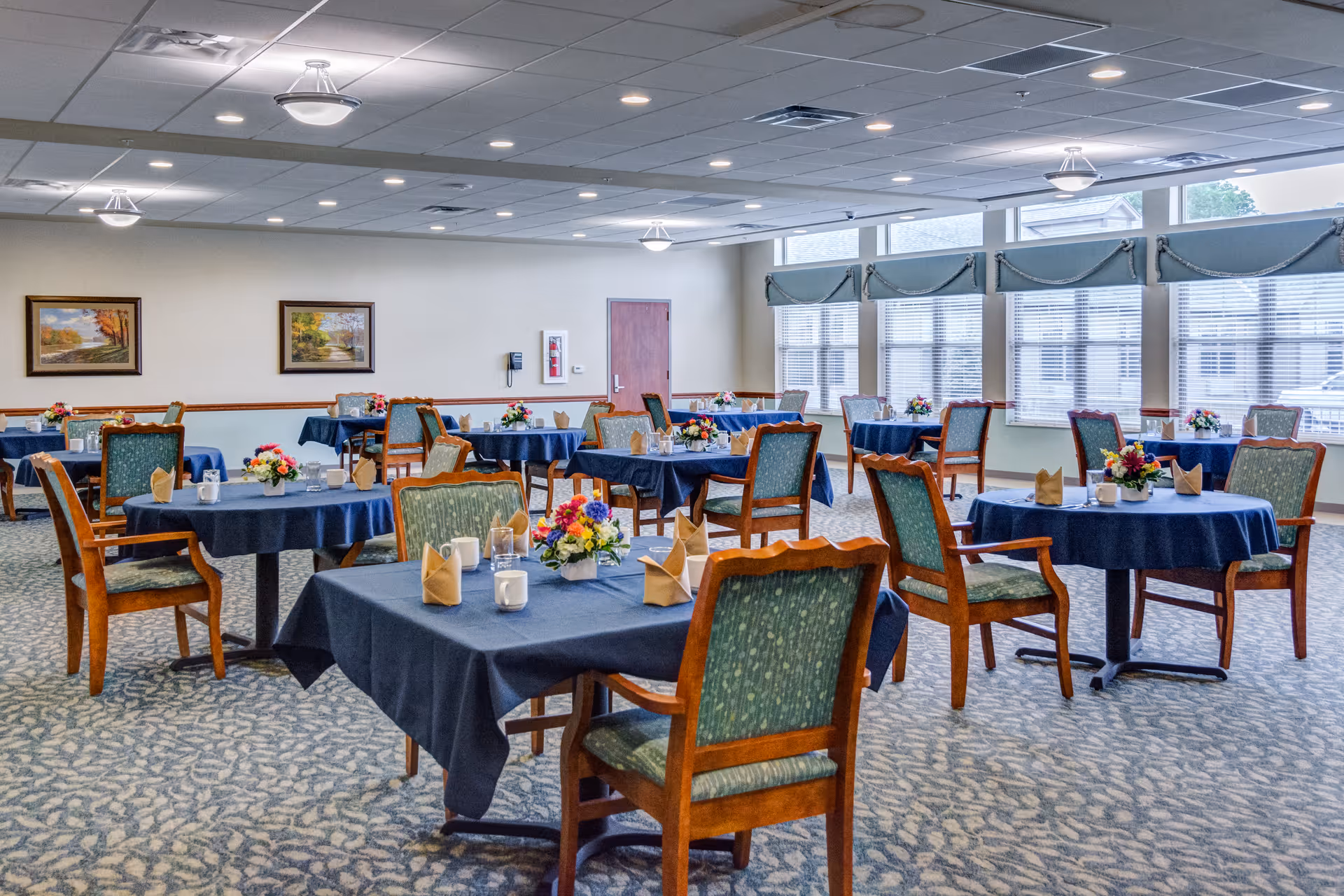 A bright dining room with round and rectangular tables covered in blue tablecloths, wooden chairs, floral centerpieces, and folded napkins.