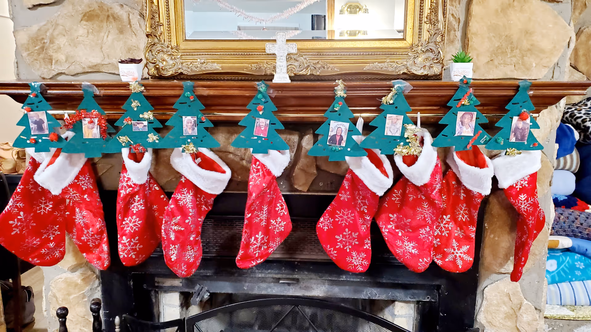 A stone fireplace with a wooden mantel decorated for Christmas. Eight red stockings with white snowflake patterns hang from the mantel, each topped with a green paper Christmas tree cutout adorned with small decorations and a photo of a person. Above the mantel is a large ornate gold-framed mirror and a white cross decoration.