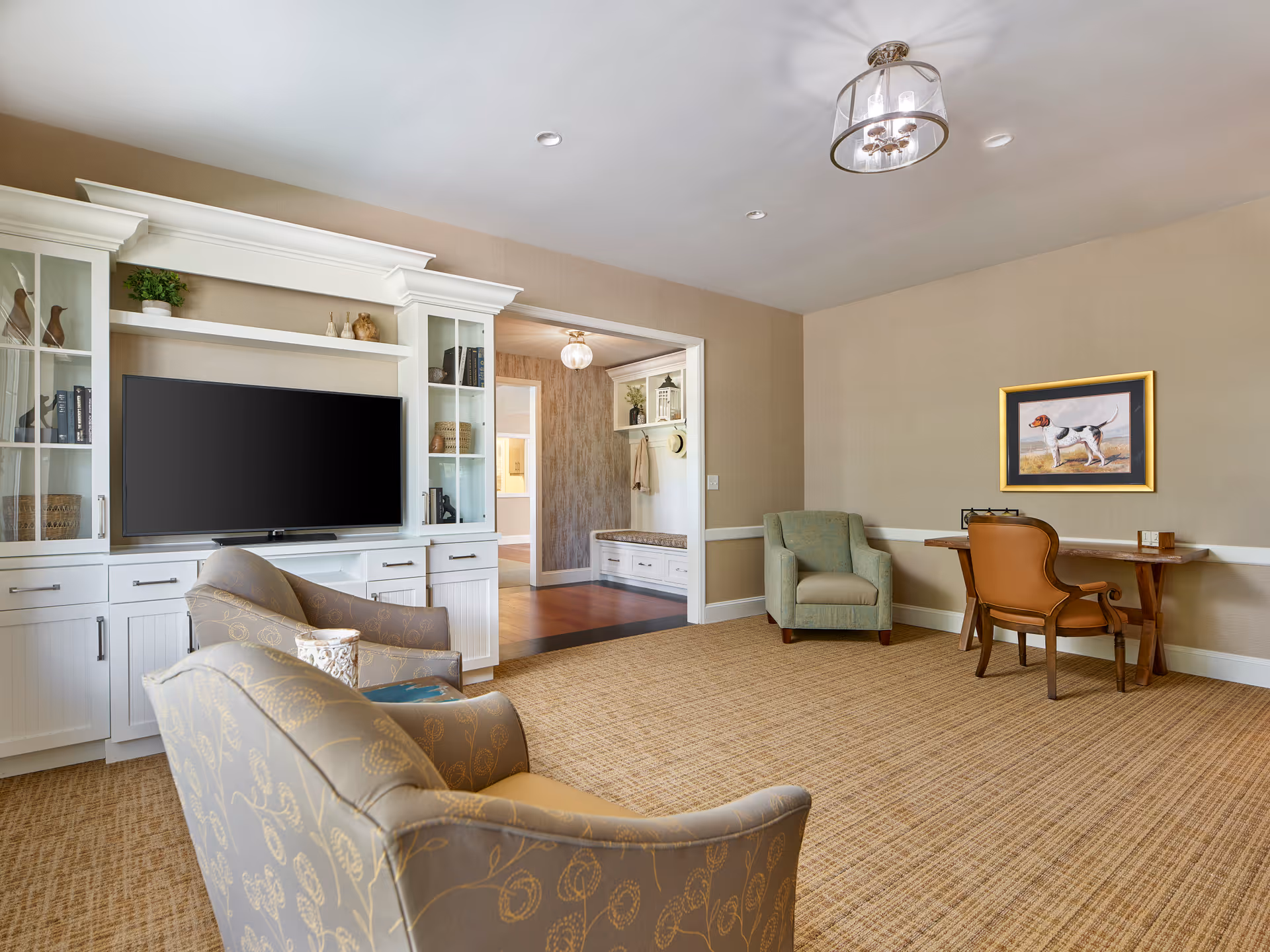 A cozy living room in a retirement community featuring a large white entertainment center with a flat-screen TV, two patterned armchairs, a green armchair, and a wooden desk with a brown leather chair. The room has beige walls, a carpeted floor, and a framed picture of a dog on the wall. There is an open doorway leading to another room with wooden flooring and a bench with hooks for coats.