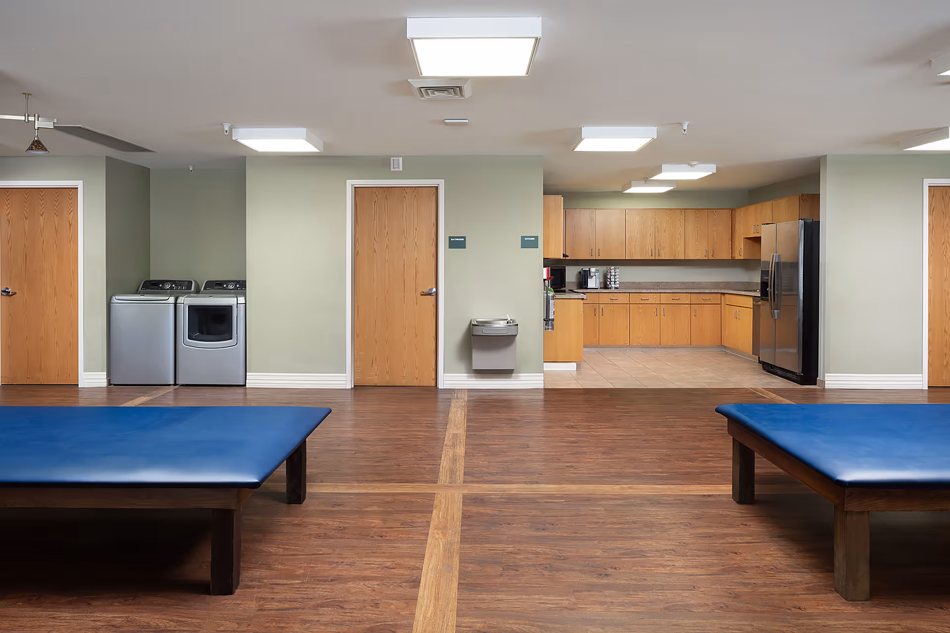 Spacious interior therapy/commons room with two blue treatment tables, wood flooring, a kitchenette, and a washer and dryer.