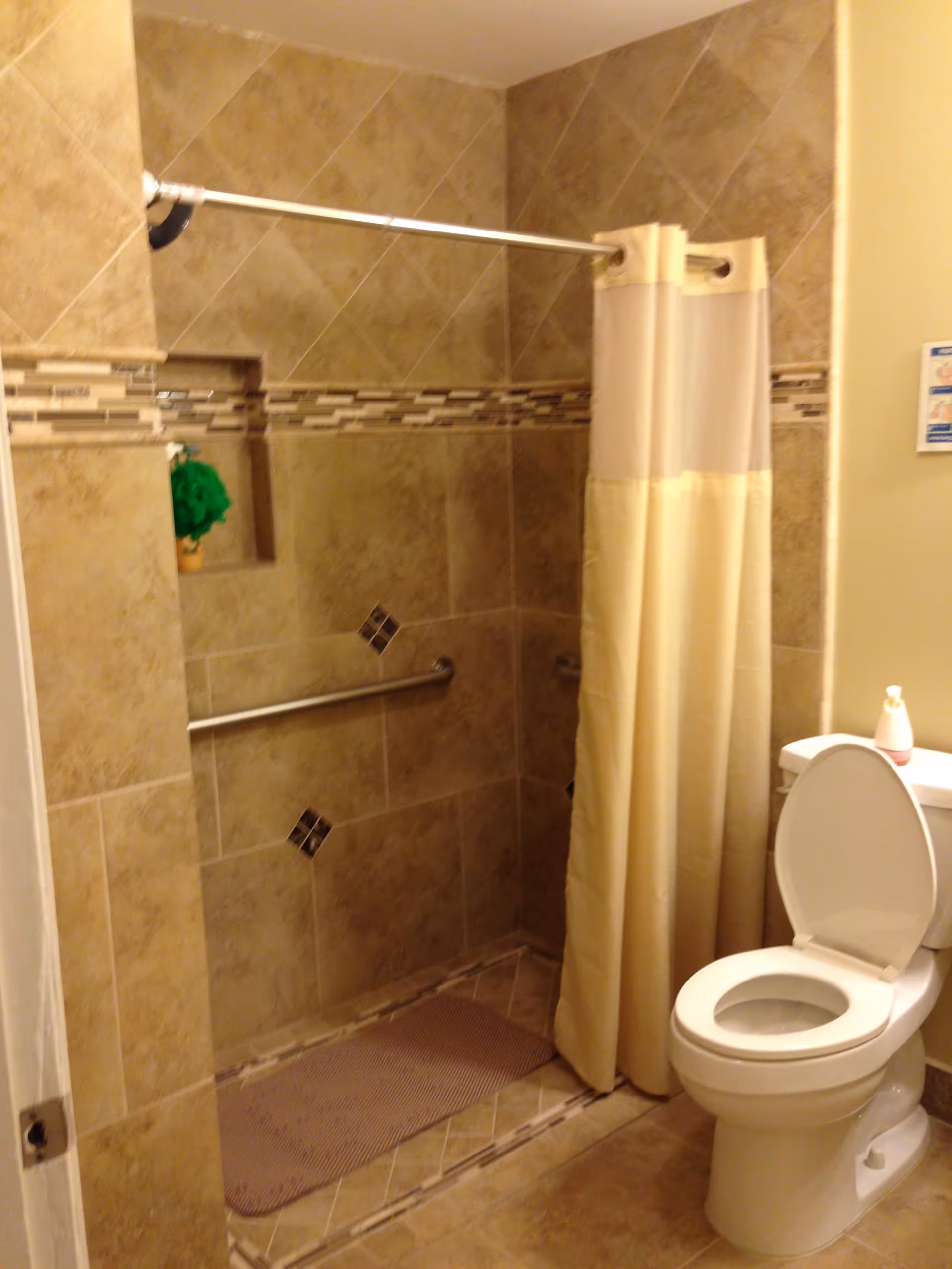 A bathroom with beige tiled walls and floor, featuring a walk-in shower with a grab bar and a beige shower curtain. There is a white toilet with the lid open next to the shower, and a small green plant is placed in a recessed shelf within the shower wall.