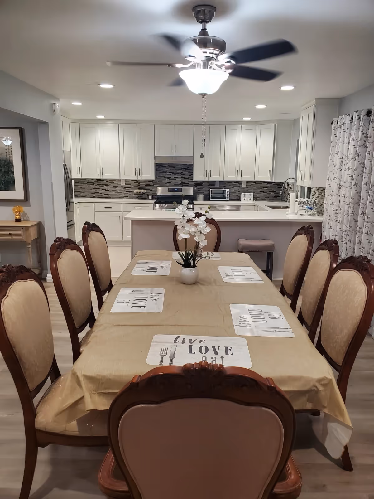 A dining room with a long table covered with a beige tablecloth and six upholstered wooden chairs. The table has placemats with the words 'live LOVE eat' and a white flower centerpiece. In the background, there is a modern kitchen with white cabinets, a tiled backsplash, stainless steel appliances, and a ceiling fan with lights above.