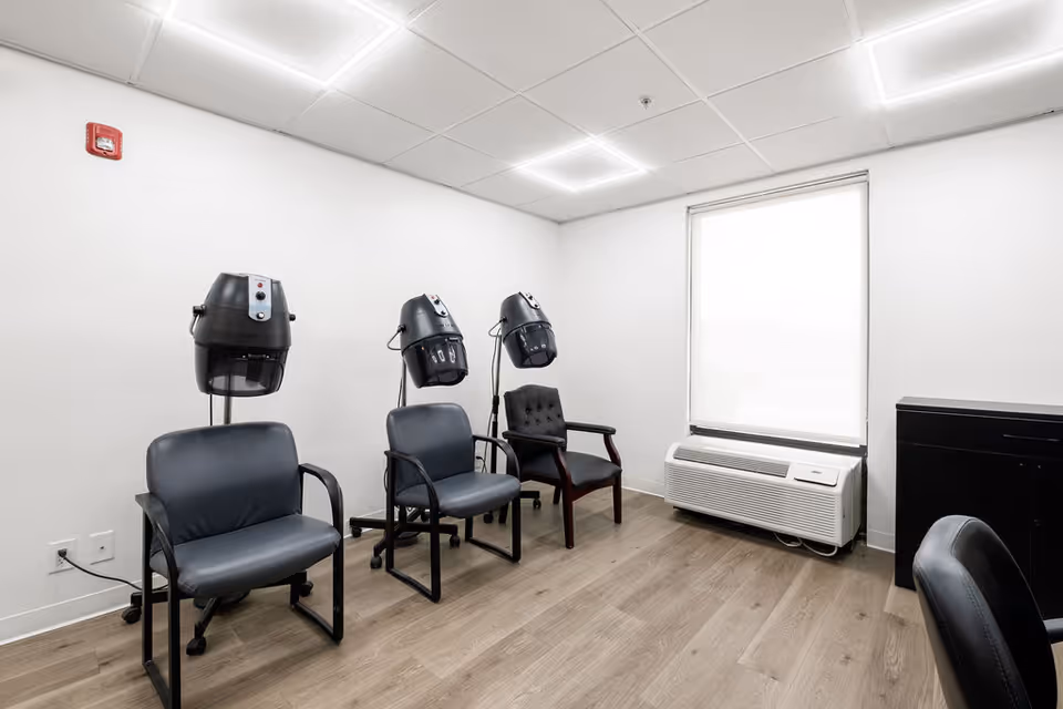 A small room with three salon chairs, each positioned under a hair dryer hood. The room has white walls, a window with a white blind, a wall-mounted air conditioning unit below the window, and a wooden floor. There is a black cabinet on the right side and a fire alarm on the wall.