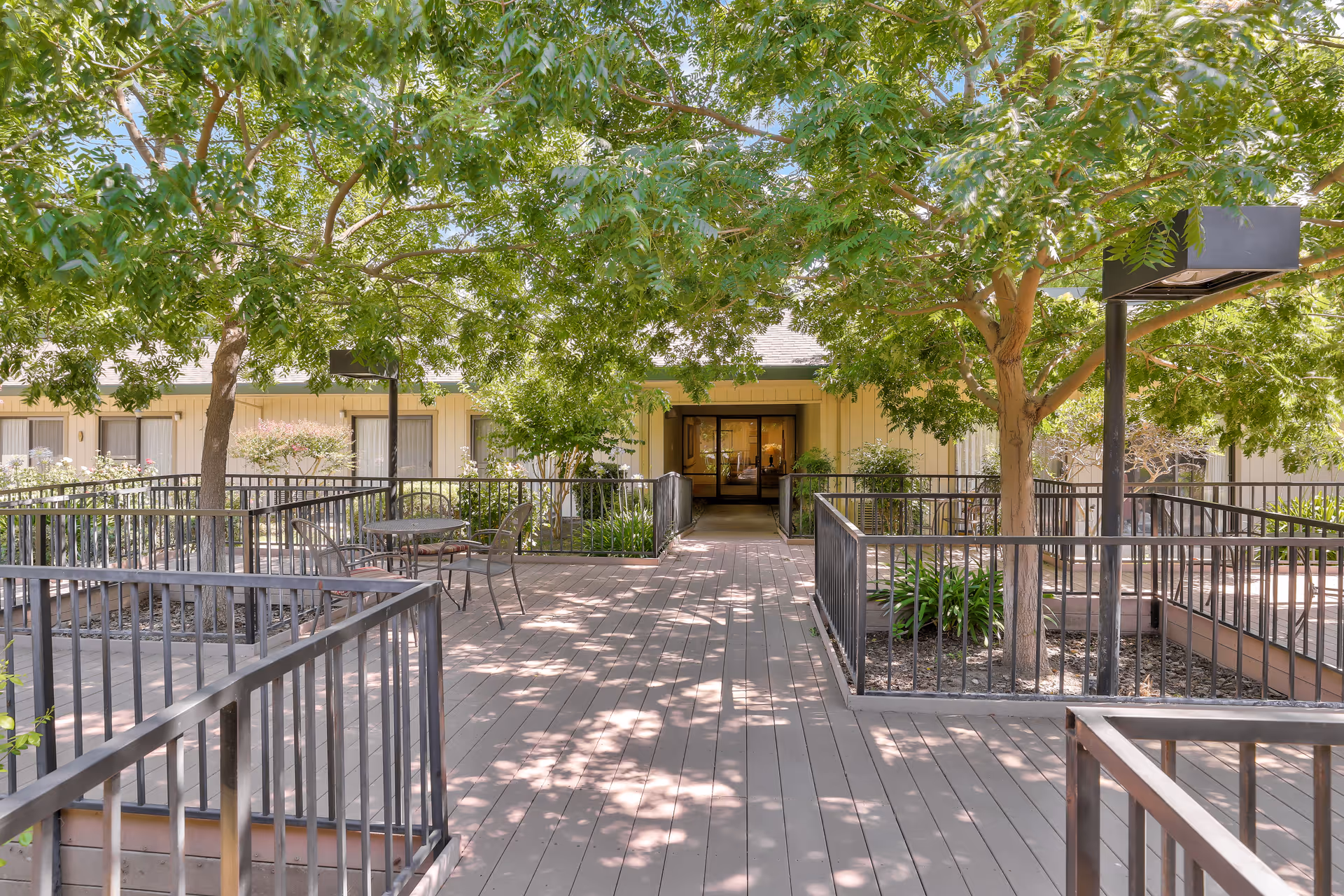 Outdoor patio area at Park Merced Assisted Living with wooden decking, metal railings, several trees providing shade, and a few metal chairs and tables. The building entrance is visible in the background.