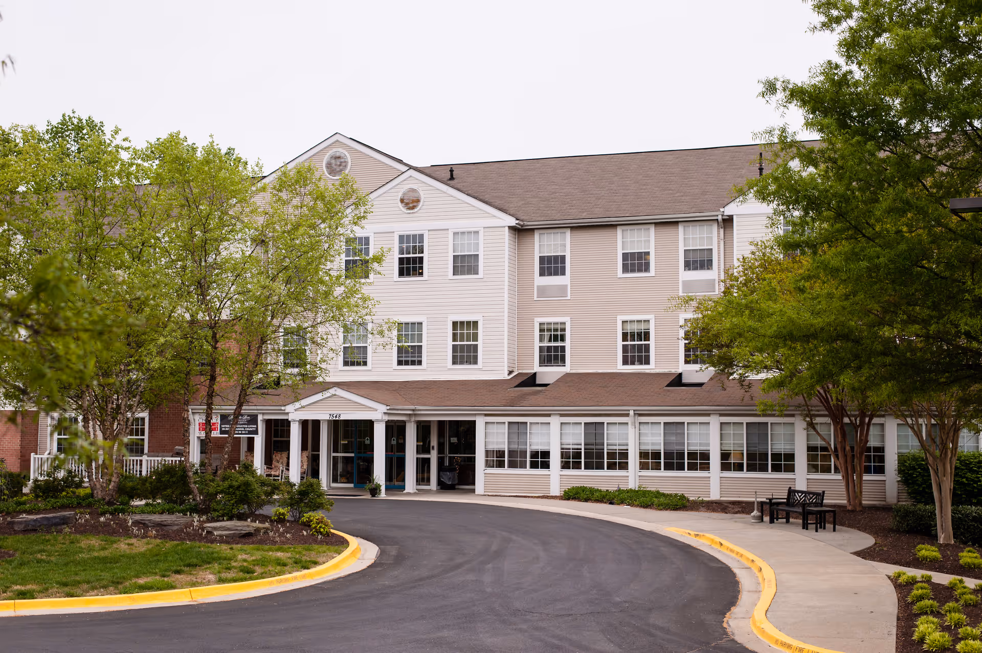 Front exterior view of a three-story senior living facility named Morningside House of Friendship, with a curved driveway, trees, and landscaped greenery around the entrance.